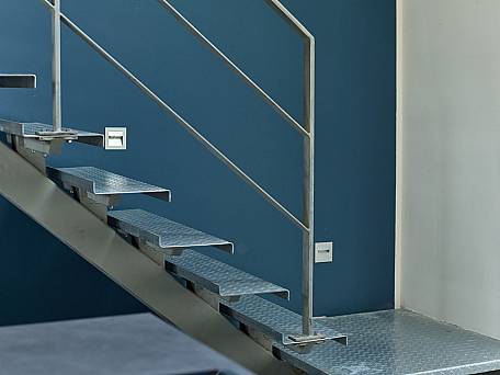 Einholmtreppe mit Stufen aus gekantetem Tränenblech, Flachstahlgeländer, Wandeinbauleuchten für Licht auf Stufen.